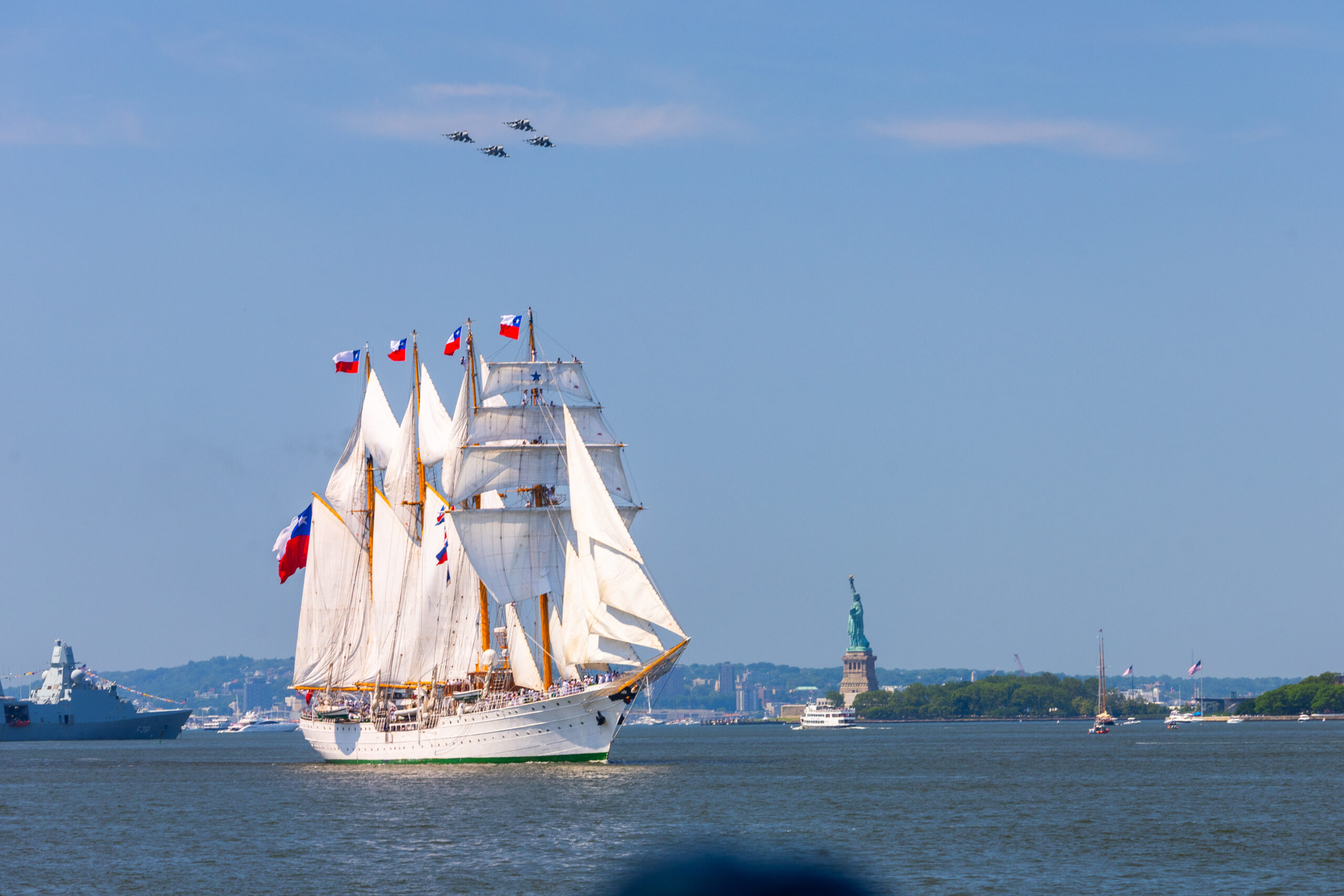 Tall ships sail into New York Harbor with aerial display overhead and Statue of Liberty in background