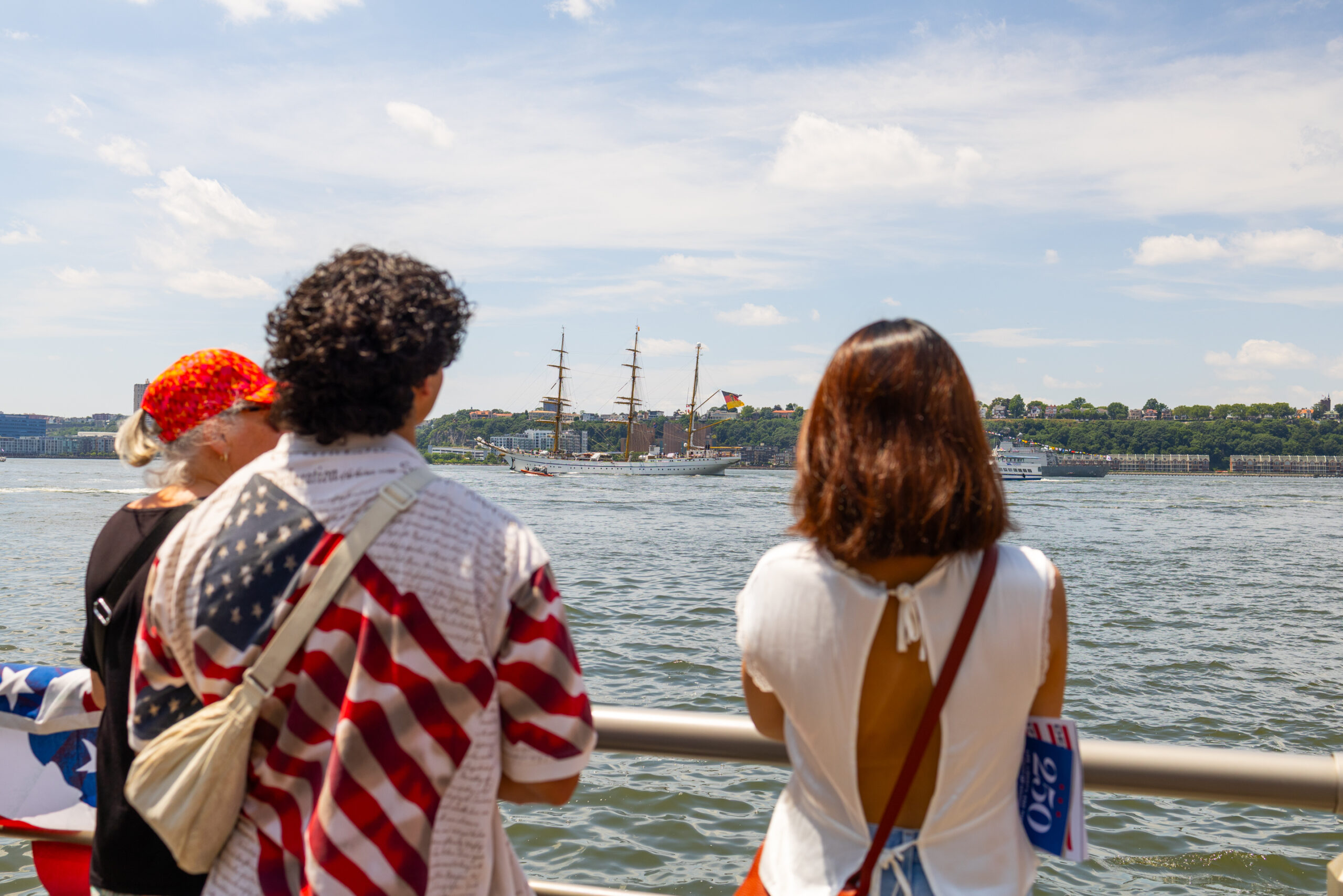 Three people stand in front of railings in front of Hudson River. One is wearing a shirt with American flags.