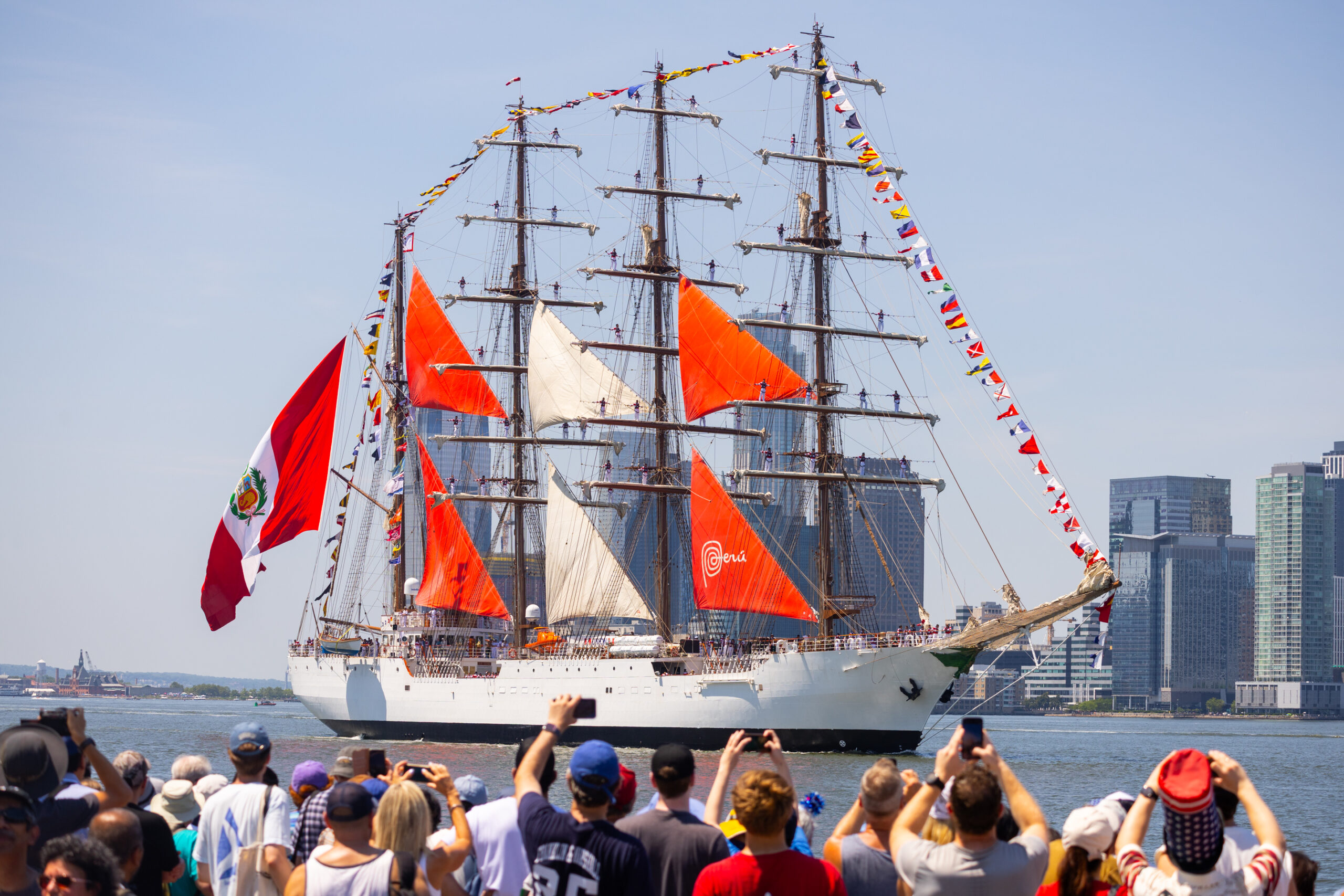 Crowds wave at tall ship with red and white sails sailing along Hudson River waterfront