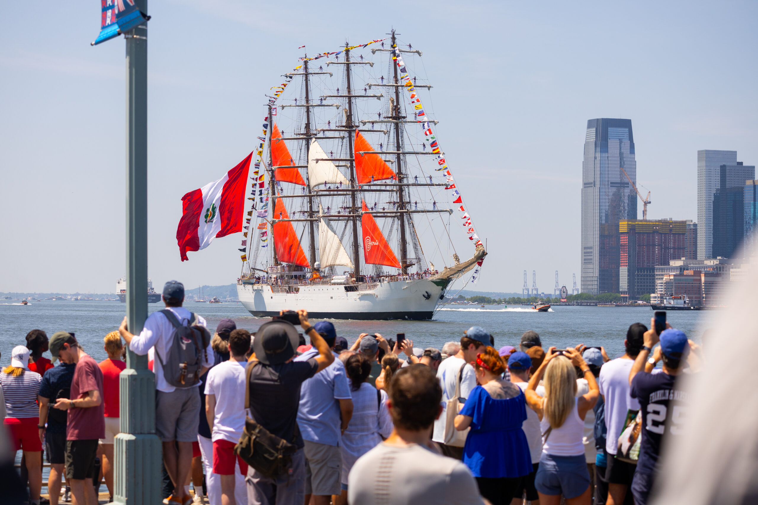 Tall ship with red and white sails sails by crowd at Hudson River Park
